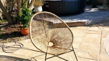 Une terrasse avec un fauteuil en osier, une table ornée d’un vase et d’un verre de jus de fruits, et un jacuzzi en arrière-plan.