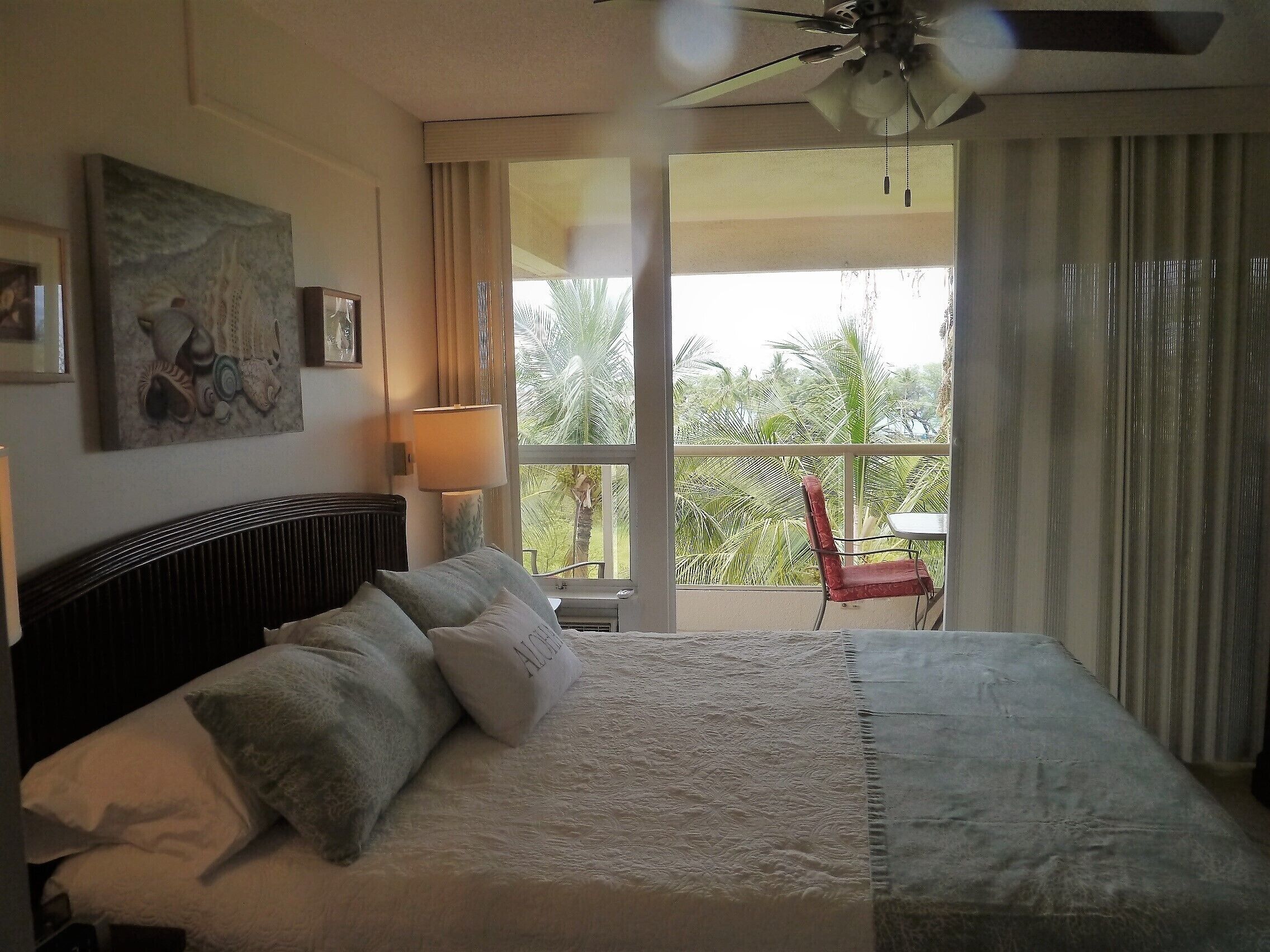A bedroom with a bed, a ceiling fan, a window with curtains, and a view of palm trees outside.