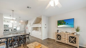 A modern kitchen with white cabinets, a dining area with a wooden table, and a living space with a TV and a plant.