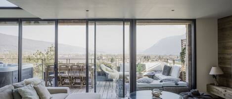 A modern living room with a large glass wall, a sofa, a coffee table, and a view of a mountainous landscape.