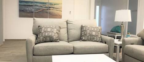 A living room with a beige sofa, patterned cushions, a white coffee table with decorative items, and a painting of a beach scene on the wall.
