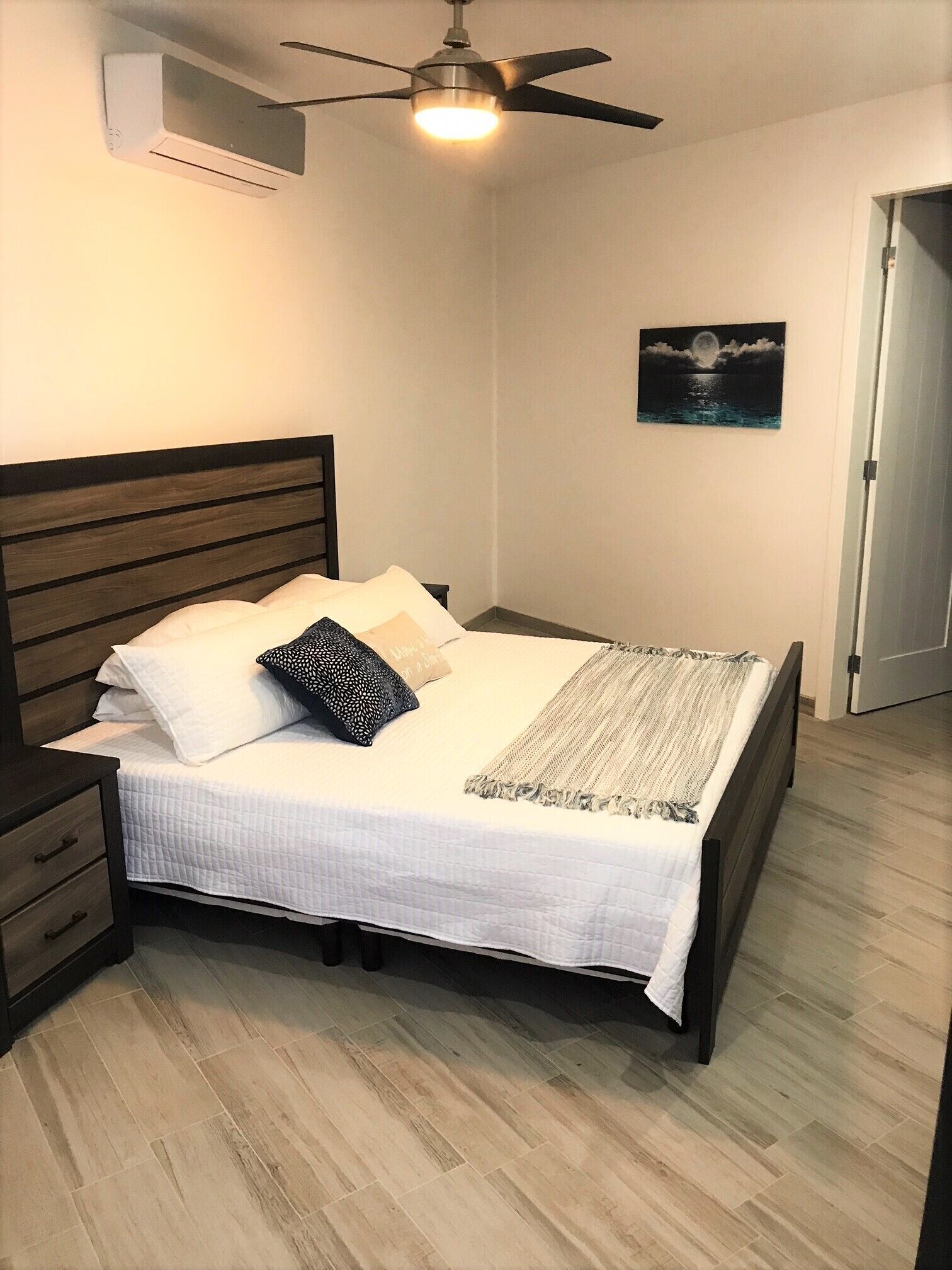 Quarto com cama, ventoinha no teto, ar condicionado, mesa de cabeceira, gaveta, piso de madeira e porta.