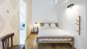 A bedroom with a wooden bed, a chair, a nightstand, a wall-mounted lamp, and a washing machine in the background. - Odorico - En centre-ville, proche gare (Rennes)
