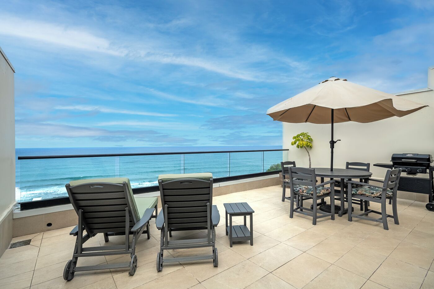 A balcony with a view of the ocean, a table with chairs, a grill, and a potted plant.