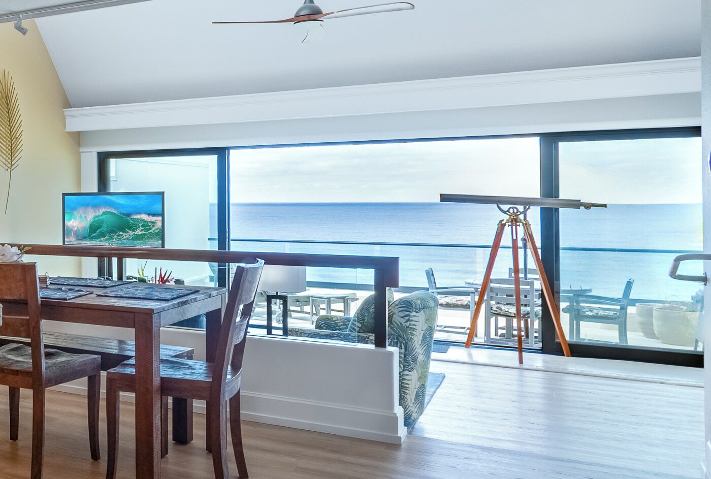 A modern room with a large window overlooking the sea, a dining area with wooden furniture, and a telescope on the windowsill.