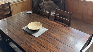 Un comedor con mesa de madera, sillas y una butaca colgante. Se observan ventanas con persianas, una pared de piedra y un techo con vigas de madera.