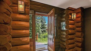 Ein Blockhaus-Interieur mit einer Holztür, die nach draußen führt, Steinböden und einem Fenster mit Blick auf Grünflächen.
