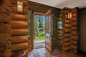 A log cabin interior with a wooden door leading outside, stone flooring, and a window with a view of greenery.