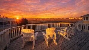Ett trädäck med Adirondack-stolar och ett bord, med utsikt över en solnedgång över en strand och avlägsna byggnader.