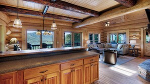 Interior espaçoso de uma cabana de toras, com ampla área de cozinha, móveis de madeira e grandes janelas que oferecem vista para o exterior.