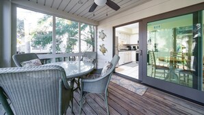 Un patio cubierto con mobiliario de mimbre, un ventilador de techo y amplios ventanales que ofrecen vistas del exterior.