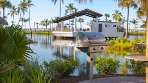 Ein Grill in Wassernähe, mit Palmen und einem modernen Haus im Hintergrund.