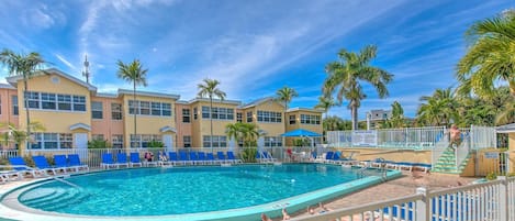 Un área de piscina con sillones reclinables, palmeras y un edificio con varias ventanas.