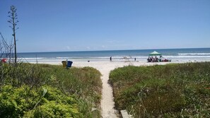 Un sentier qui longe la plage et mène au rivage, avec une tente verte et des personnes se baignant en arrière-plan.