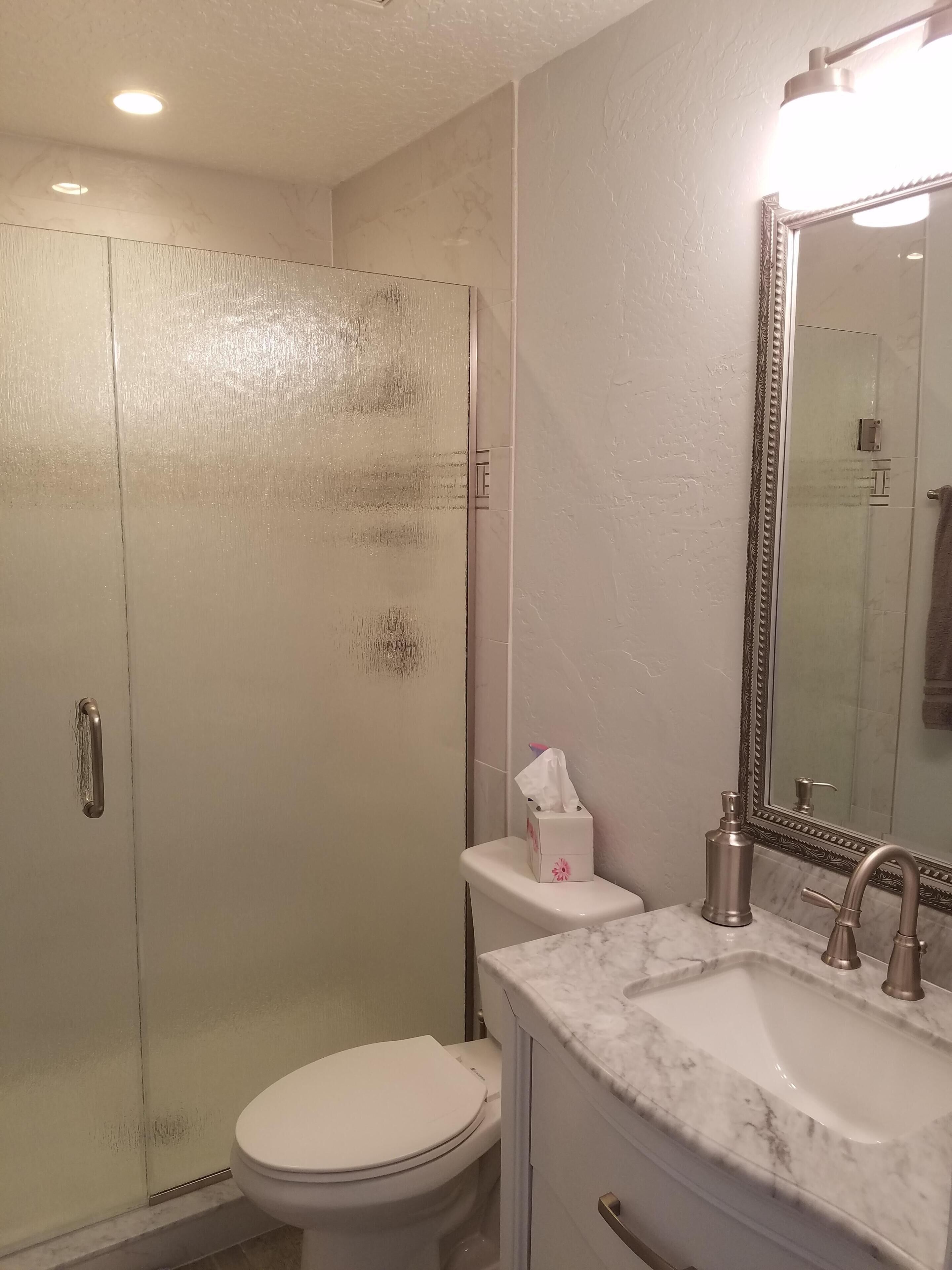 Un baño con inodoro, lavamanos con encimera de mármol, ducha con puerta de vidrio y espejo con marco decorativo.