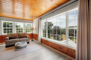 A room with a wooden ceiling, large windows, a sofa, a lamp, and a rug.