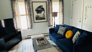 A living room with a dark blue sofa, patterned cushions, a coffee table, a flat-screen TV, and a framed artwork on the wall.