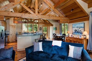 A spacious living area with a dark blue sofa, wooden ceiling beams, a kitchen island, and large windows.