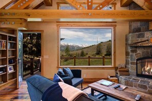 A cozy living room with a fireplace, a large window with a mountain view, a bookshelf, and a sofa.