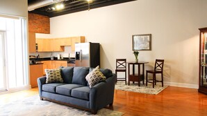 A modern living room with a gray sofa, wooden dining table, and chairs. A kitchen with wooden cabinets and appliances is visible in the background.