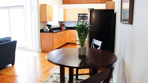 A dining area with a round table, chairs, a potted plant, and a kitchen in the background.
