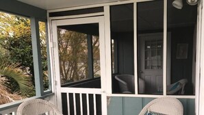Une véranda avec deux fauteuils en osier, dont l’un est agrémenté d’un coussin bleu, et une porte donnant sur une pièce intérieure.