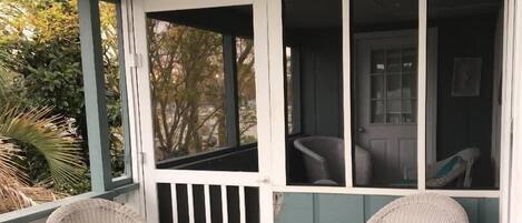 Un porche con dos sillones de mimbre, uno con cojín azul, y una puerta que da a una habitación interior.