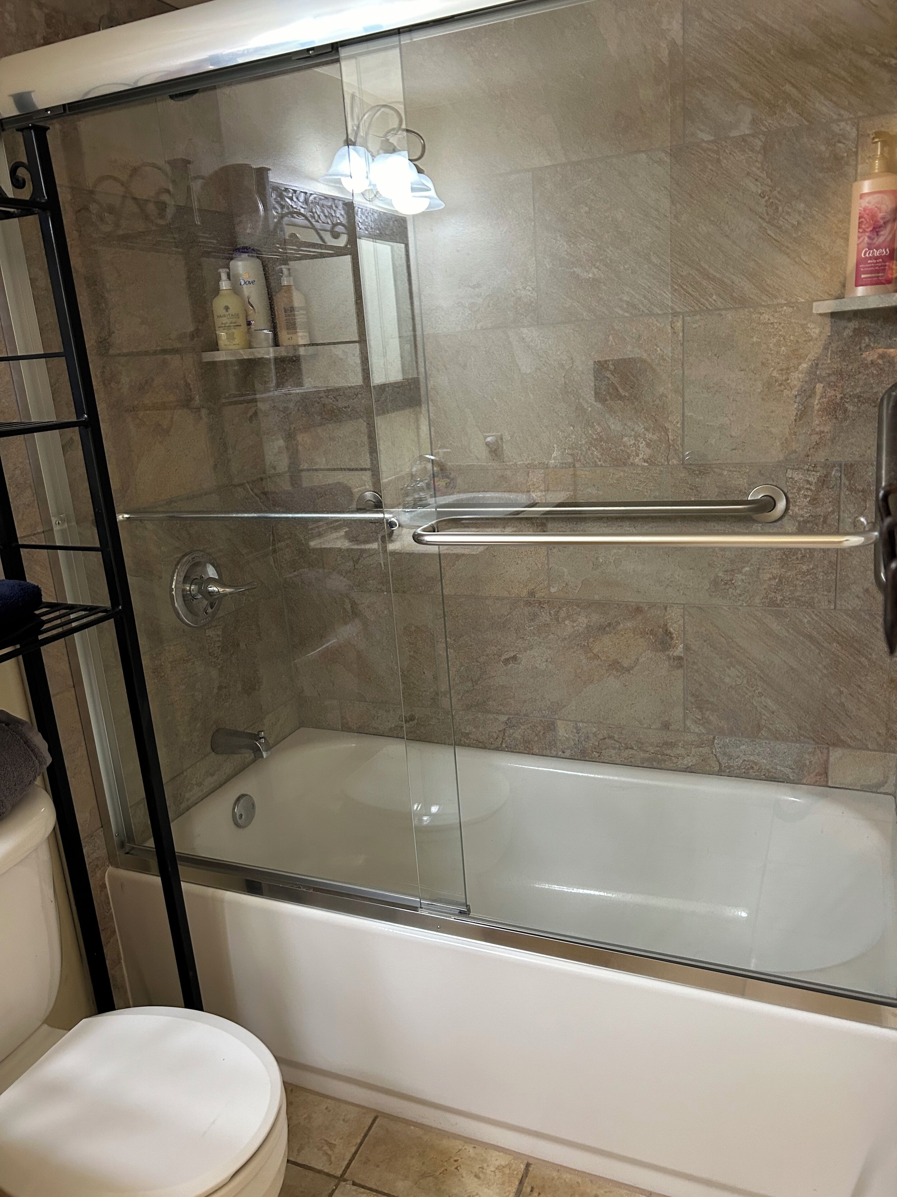 Une douche fermée par des parois en verre, dotée d’une barre d’appui, une baignoire blanche et une étagère contenant des articles de toilette.