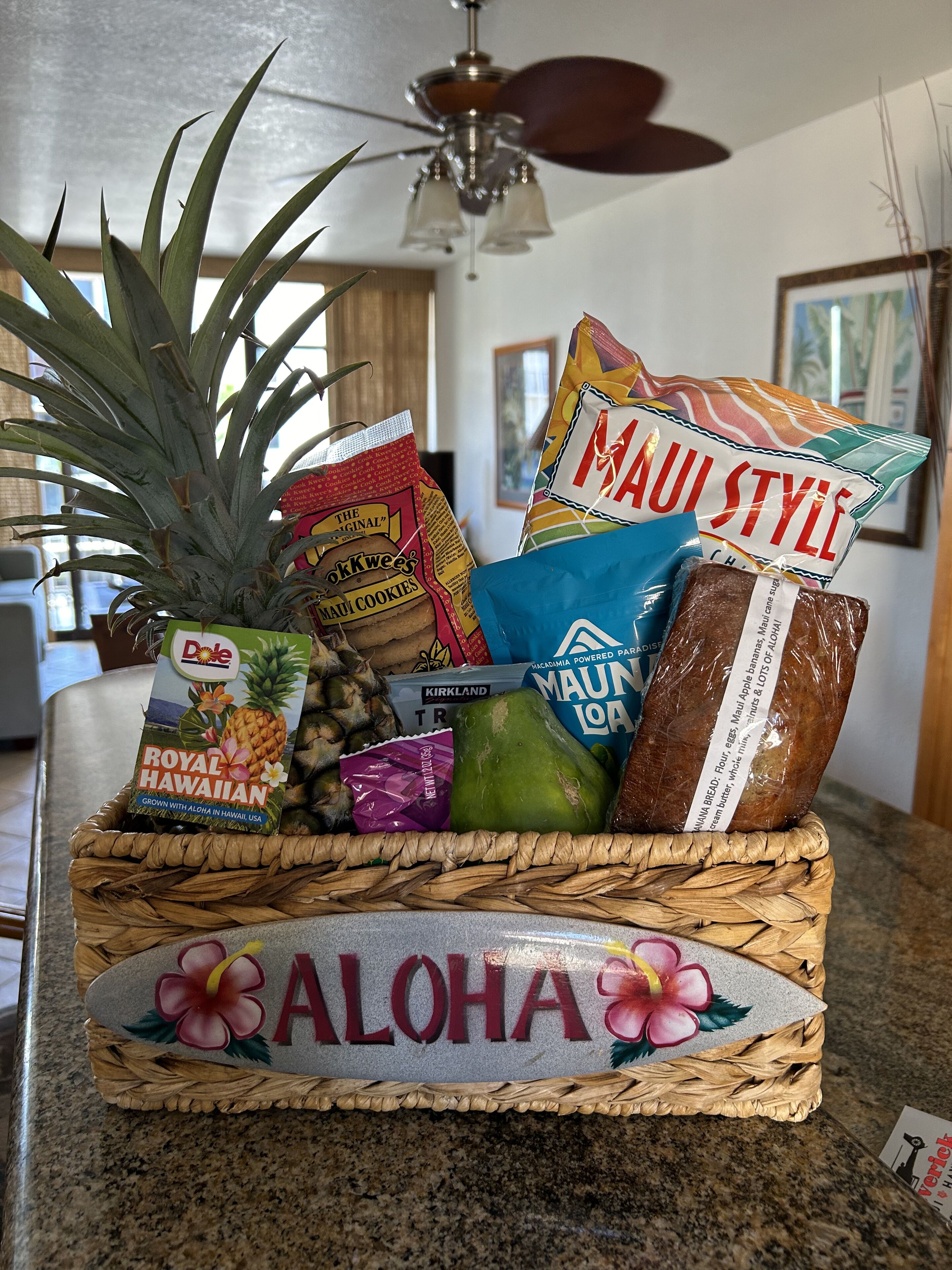 Una cesta tejida llena de objetos con motivos hawaianos, como una piña, galletas y pan, colocada sobre una encimera de cocina.