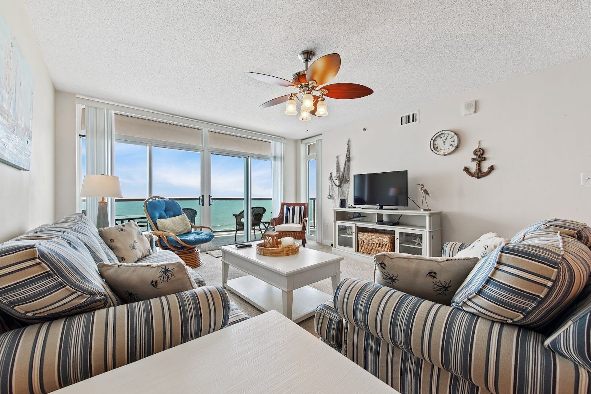 A living room with striped sofas, a coffee table, a ceiling fan, a TV, and a view of the ocean through large windows.