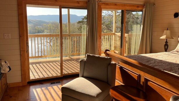 A cozy room with a bed, a chair, a small table, and a balcony with a view of a lake and mountains. - Authentic Mountain Log Cabin Perched On A Cliff Side Above The Shenandoah River (Front Royal)