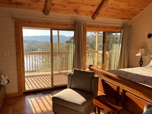 Una habitación acogedora con cama, sillón, mesita y balcón con vista a un lago y montañas.