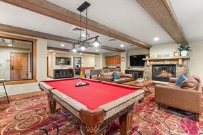 A pool table with balls, a sofa, a fireplace, and a television in a room with wooden beams and a carpeted floor.