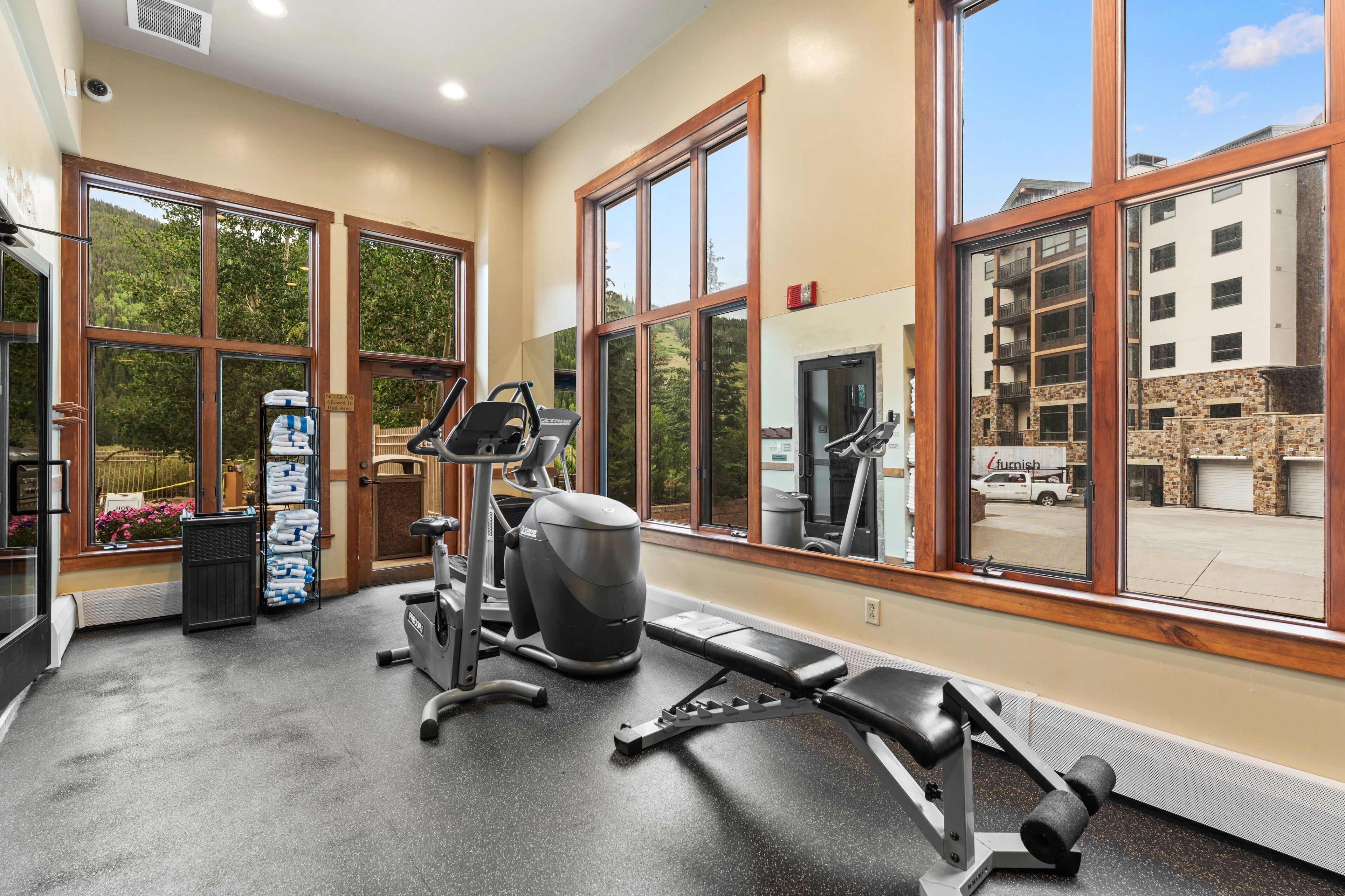 Ett gym med träningsutrustning, stora fönster och utsikt över en byggnad utanför.