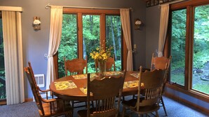 A dining area with a round wooden table, six wooden chairs, and a vase of yellow flowers. Large windows with curtains provide views of a forested area. - RELAX & GETAWAY IN A FULY FURNISHED CABIN IN THE BLUE RIDGE SMOKEY MOUNTAINS.  (Maggie Valley)