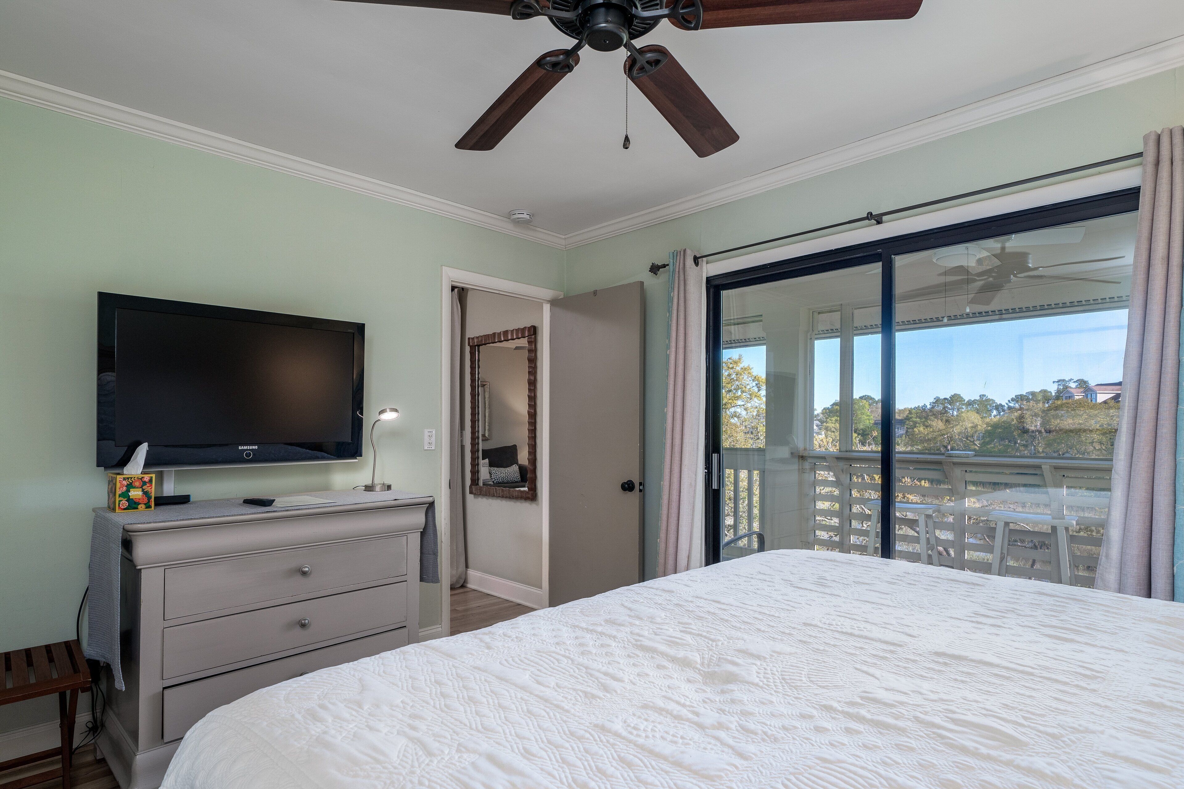 A bedroom with a bed, a dresser, a television, a ceiling fan, and a sliding glass door leading to a balcony.