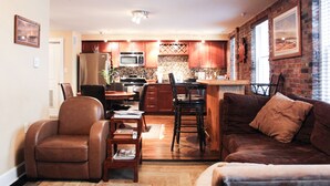 A cozy living room with a brown leather sofa and a matching armchair, a dining area with a table and chairs, and a kitchen with wooden cabinets and a brick wall. - Welcome to Derby City Loft! (Louisville)