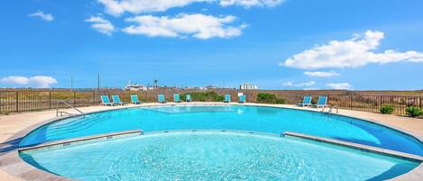 A large, curved swimming pool with clear blue water, surrounded by a low stone wall and a metal fence.