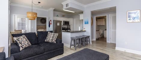 A modern living room with a dark grey sofa, a matching ottoman, and a kitchen area with white cabinets and a stainless steel refrigerator.
