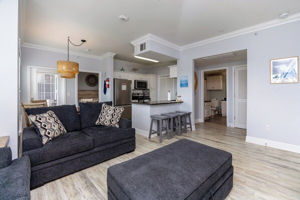 A modern living room with a dark grey sofa, a matching ottoman, and a kitchen area with white cabinets and a stainless steel refrigerator. - First-Floor Condo | Steps to Beach & Private Patio (Corpus Christi)