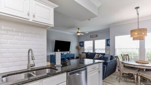 Cocina moderna con armarios blancos, un lavamanos de acero inoxidable y lavavajillas. Hay un comedor con mesa y sillas de madera, un sofá azul y un ventilador de techo.