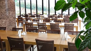 En rummelig restaurant med træborde og stole, store vinduer med udsigt til grønne omgivelser.