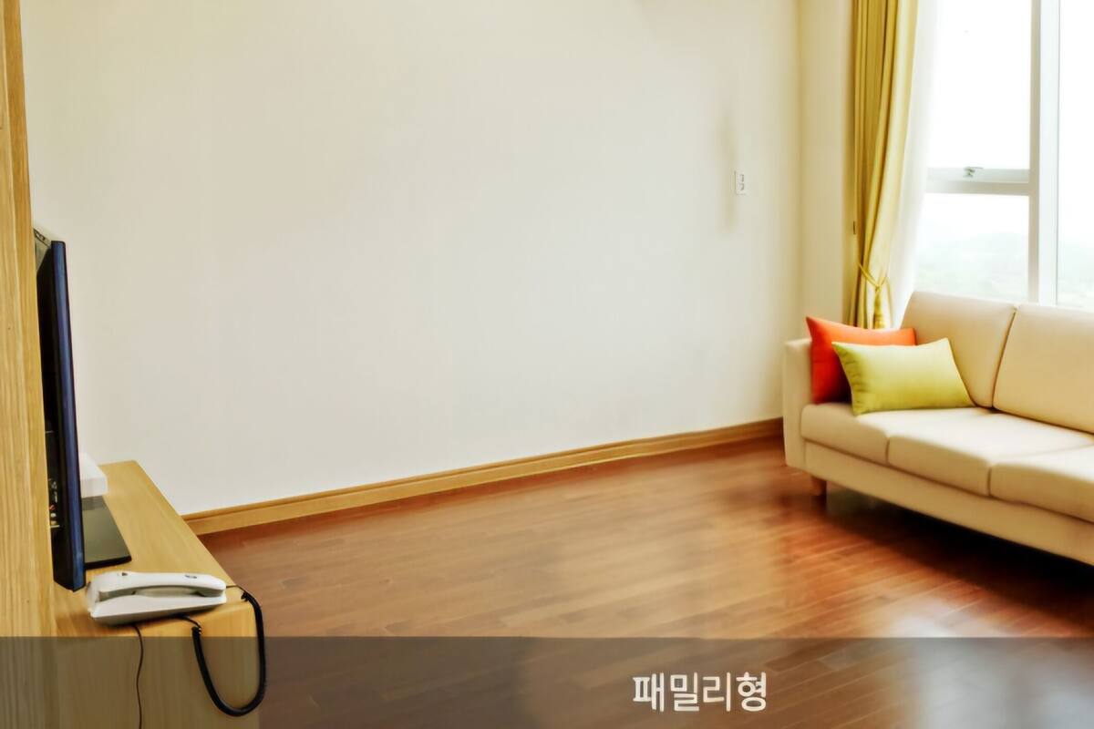 A room with a beige sofa, a flat-screen TV on a wooden table, and a window with curtains.