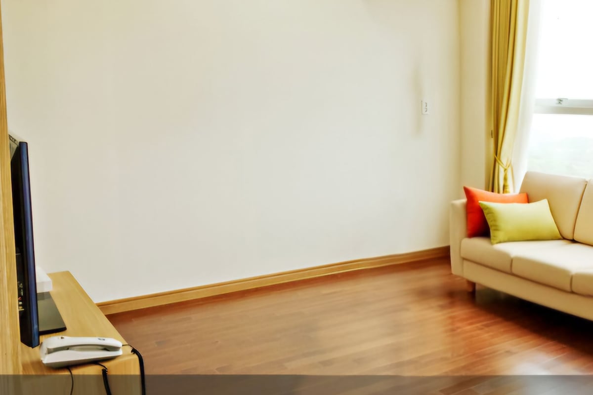Een kamer met een beige bank, een flatscreen televisie op een houten tafel en een raam met gordijnen.