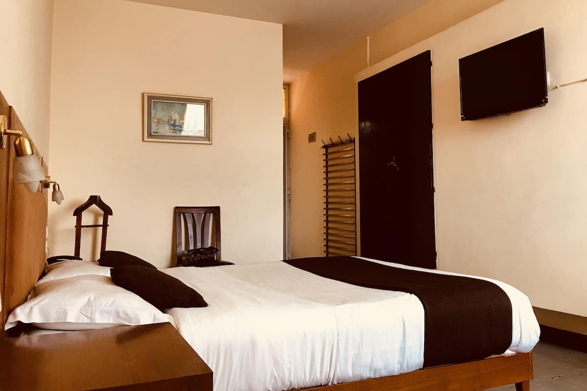 Une chambre à coucher avec un lit, une tête de lit en bois, une table de chevet, un tableau encadré au mur, une fenêtre avec des stores et un téléviseur fixé au mur.