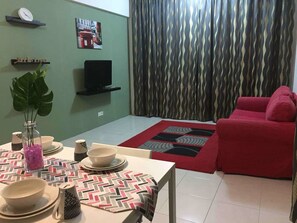 Una sala de estar con un sofá rojo, un televisor sobre un mueble, una mesa de comedor con platos y tazones, y una alfombra estampada.