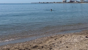 Una playa con agua cristalina, una persona nadando y estructuras industriales al fondo.