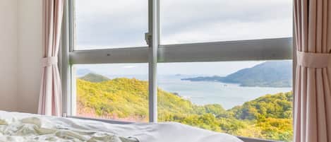 A bedroom with a large window offering a view of a mountainous landscape and a body of water.