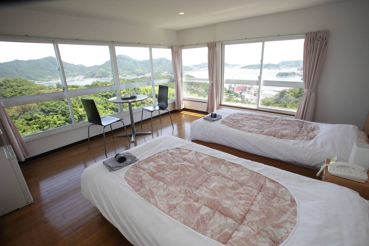 Camera d'albergo con due letti, un tavolino, una sedia e vista su un paesaggio montuoso attraverso ampie finestre.