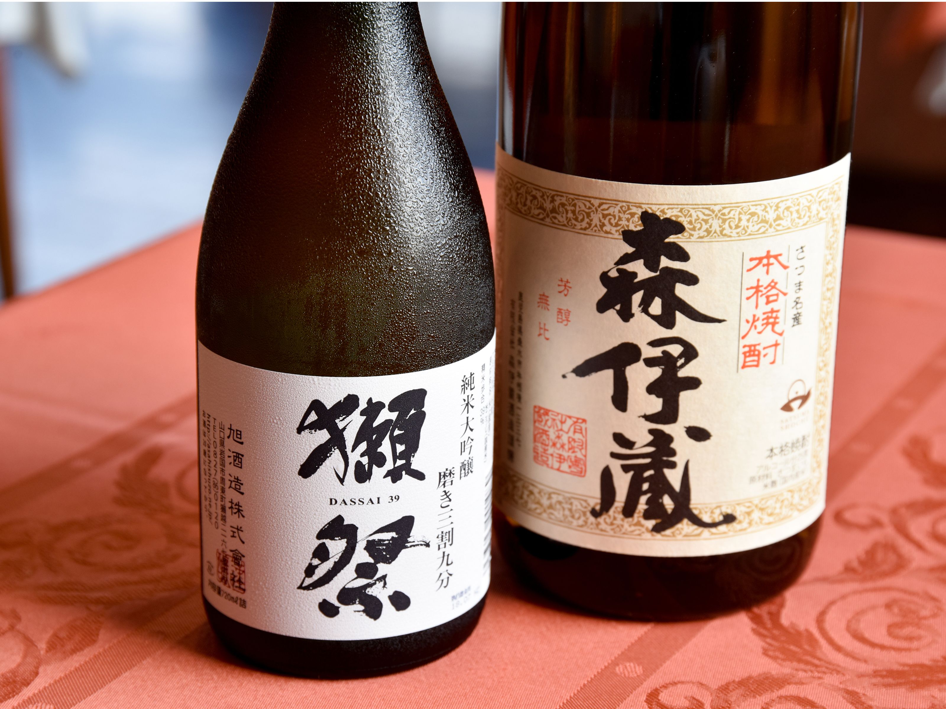 Dos botellas de sake japonés con etiquetas, sobre una mesa con mantel estampado.